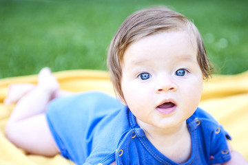 Newborn girl with blue eyes in the beautiful park outdoors, lies on a blanket wearing blue babybody and looking into camera

