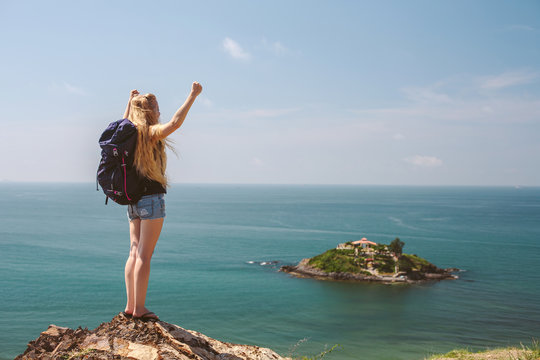 Happy woman staying on the mountain cliff, raising arms towards beautiful island on blue ocean