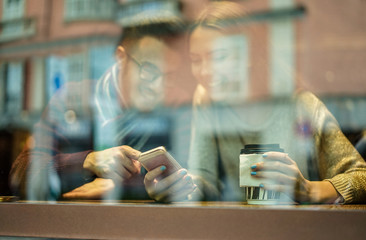 Young happy couple in coffee bar window watching mobile phone