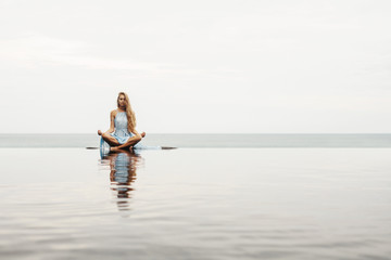 Beautiful female in blue dress sitting near sea beach in lotus pose, relax and meditate. Water...