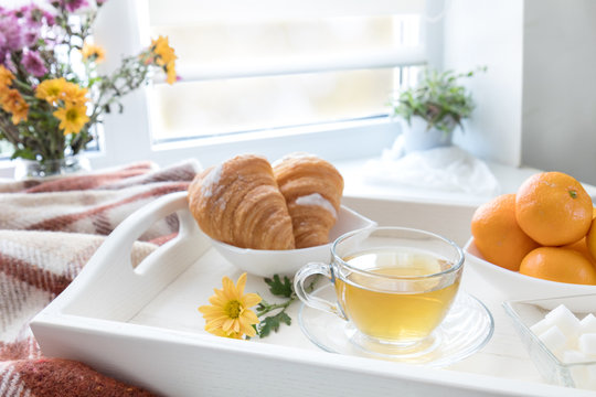 Cup of tea, croissant, chrysanthemum flowers and tangerines in tray on the windowsill. Cozy home concept. Coloring and processing photo