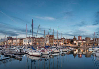 Fototapeta na wymiar Yatchs and pier in leisure port on maritime fishing district of Gijon, Spain, Europe. Beautiful reflection on calm sea water of boats, buildings, sky at dusk at touristic cultural travel destination.