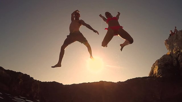 SLOW MOTION CLOSE UP LOW ANGLE VIEW: Two happy playful friends jumping off sharp rocky ledge into stunning ocean with hands raised at magical pinky summer sunset. Water drops splashing and sprinkling
