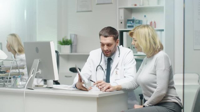  Professional Doctor Consults Mid Adult Woman. He Points out Something on the Screen to Her. Nurse Puts Documents on Doctor's Table. Shot on RED Cinema Camera in 4K (UHD).
