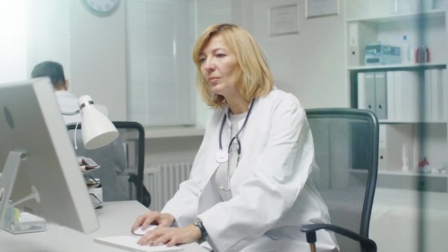 In the Doctor's Office. Mid Adult Female Doctor Works at Her Desktop Computer. Her Assistent Stands Up and Leaves the Office. Shot on RED Cinema Camera in 4K (UHD).