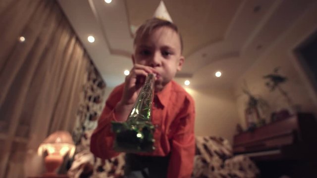 Little boy in Birthday cap holding sparkler and blowing whistle at camera in modern room.