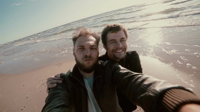 Two young goofy guys with beards are standing on a sandy beach trying to take a selfie.