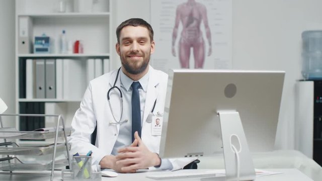  
Male Doctor is Working at His Desk. He Interrupts His Work, Folds His Hands and Smiles at the Camera. Shot on RED Cinema Camera in 4K (UHD).
