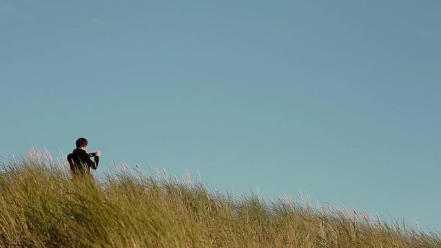 A dark-haired man is standing in tall grass taking a picture on his phone on a windy day