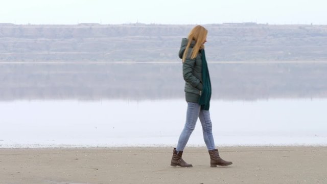 Young beautiful nice smart girl walking on sandy beach and smiling. Quiet weather, calm water, fog, mist and haze over water. Beach is reflected on water surface. Fantastic landscape film or movie.