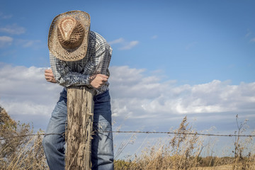 horizontal image of a caucasian cowboy standing on a barbwire fence leaning over a wood post looking down under a bright blue sky in summer time.