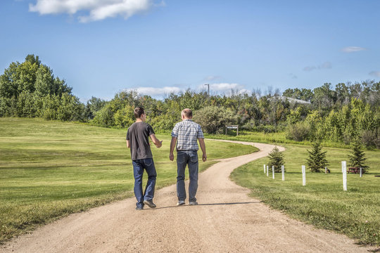 horizontal image of a caucasian father and teenage son spending quality time together talking and walking down a gravel  gravel path with lots of green grass and trees in the summer time.