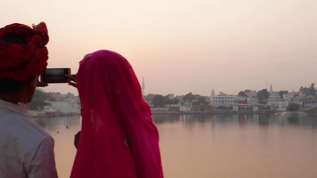 Pan left to couple in Indian dress taking photos of sunset at a holy lake in Rajasthan 