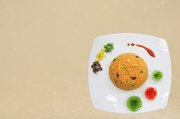 Orange fried rice decorate with vegetable such as currant, corn, peas, Japanese cucumber carving and tomato carving. The white dish on sand.