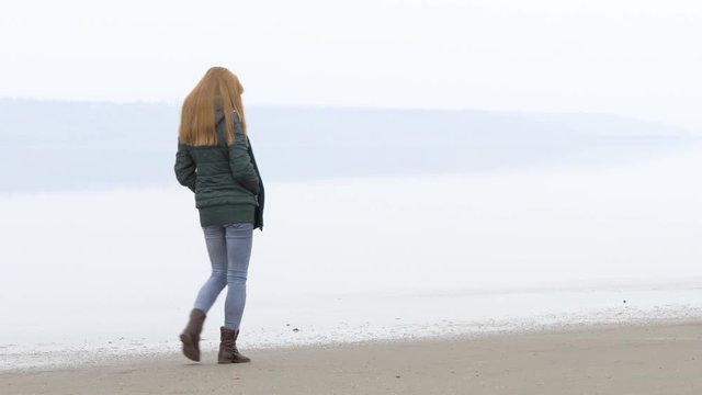 Young beautiful nice smart girl walking away on sandy beach. Quiet weather, calm water, fog, mist and haze over water. Beach is reflected on water surface. Fantastic landscape movie. Close shot