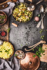 Vegetarian dishes with yellow rice,  cabbage and mushrooms in cooking pots on dark rustic country kitchen table with spoons, top view, frame