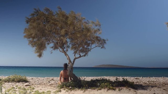 Woman sitting in the shade of a cypress tree at the beach in the summer, looking at the blue sea.