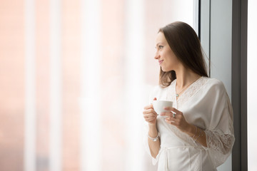 Young smiling woman in lace night gown relaxing at cozy home, feeling happy, resting in the morning, looking through the window with a cup of drink, challenge of a new day. Copy space