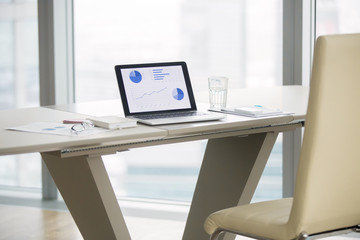 Laptop with graphs on screen on white modern office desk of business analyst, revealing business progress, seeking technology solutions, vacant place. Luxury office interior. Business success concept