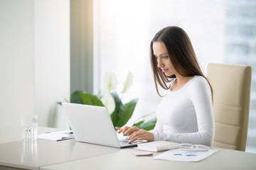 Young woman working at the modern office desk with a laptop, busy with distance learning, tutoring, making online surveys. Lady boss at her workplace. Business success concept