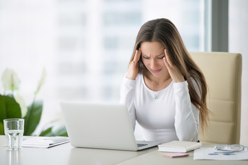 Young frustrated woman working at office desk in front of laptop suffering from chronic daily headaches, treatment online, appointing to a medical consultation, electromagnetic radiation, sick pay - obrazy, fototapety, plakaty