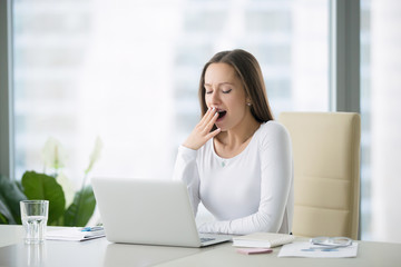 Young business woman yawning at a modern office desk in front of laptop, covering her mouth out of...