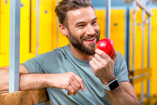 Sports Man Eating An Apple Sitting After The Training In The Locker Room Of The Gym. Healthy Natural Food For Sports Men