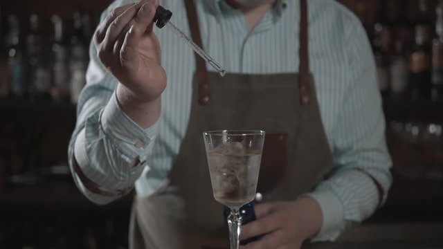 Bartender is making cocktail at bar counter, adding some bitter in the shaker