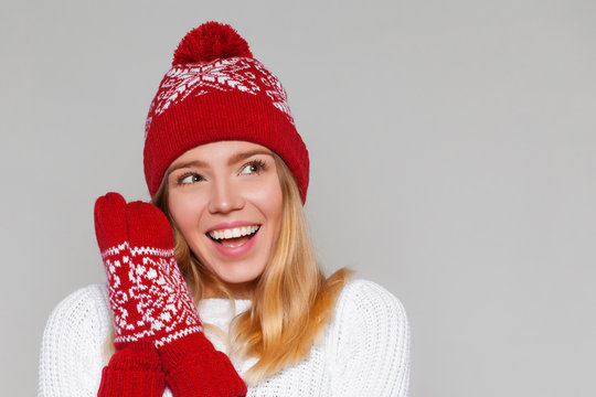 Surprised happy beautiful woman looking sideways in excitement. Christmas girl wearing knitted warm hat and mittens, isolated on gray background
