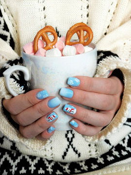 A girl wearing knitted sweater is holding a mug of hot chocolate decorated with marshmallows and pretzels