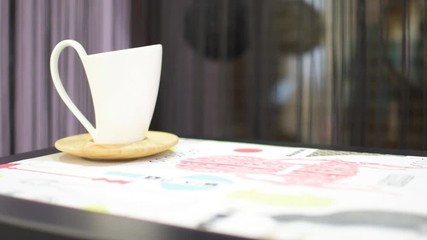 white cup of coffee on a background of black curtains on the table. In the background is an interior designer.