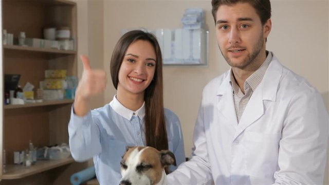 Female client showing her thumb up at the veterinarian clinic. Pretty caucasian girl approving vet clinic. Attractive young girl standing with vet doctor near the table with dog on it