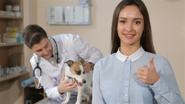 Pretty caucasian girl approving veterinarian clinic. Male veterinarian holding dog on the table. Young smiling girl showing her thumb up against background of brunette male vet with dog