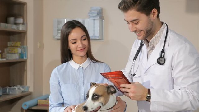 Male vet telling female dog owner about treatment for her dog. Attractive male veterinarian holding red pack of some dog treatment. Male vet doctor pointing his fingers on description of dog treatment