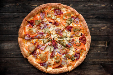 Pizza with salami, pickles, tomatoes, cheese and ham on a dark wooden background, top view