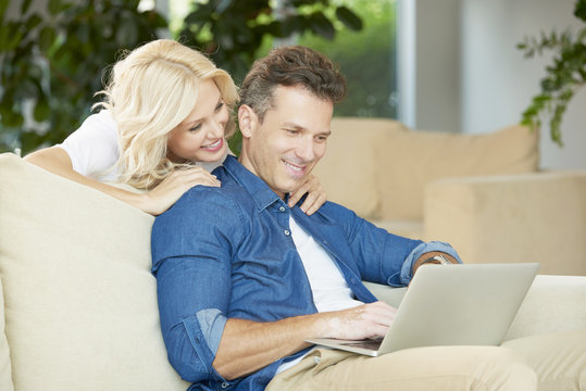 Portrait Of Middle Aged Couple Sitting With A Laptop At Home. Beautiful Blond Woman Using Laptop While Handsome Man Sitting Next To Her And Enjoy Surfing On Internet.