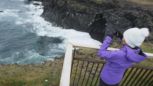 Tourist on travel taking photo with smart phone on Iceland of dramatic coast and ocean. Happy woman sightseeing taking pictures using smartphone visiting Arnarstapi, Snaefellsnes, West Iceland.