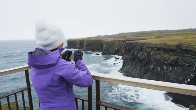 Tourist on travel taking photo with smart phone on Iceland of dramatic coast and ocean. Happy woman sightseeing taking pictures using smartphone visiting Arnarstapi, Snaefellsnes, West Iceland.
