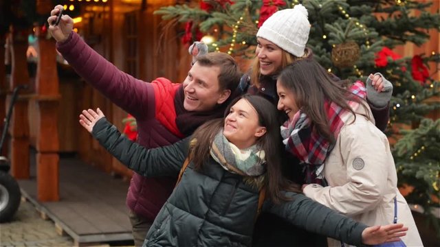 Group of Smiling Man and Women Taking Selfie Outdoors near Xmas Tree. Friends Having Fun on the Christmas Market. Merry Christmas and Happy New Year