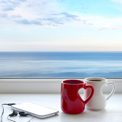 Two cups of hot tea, smartphone and headphones on a window sill. In the background sea and sky