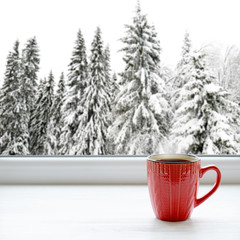 Cup of hot tea on a window sill. In the background, a beautiful winter forest in snow