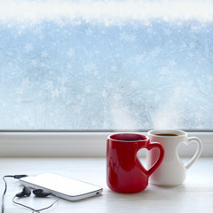Two cups of coffee, smartphone and headphones on a windowsill. In the background frosty pattern on window as a Christmas background