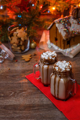 Two glasses of cocoa with marshmallows, whipped cream and chocolate syrup on wooden table and Gingerbread house, cookie jar and christmas tree with toys and garland on background.