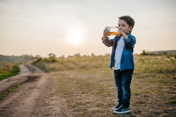 Little boy drinks juice from a bottle, stands on a rural meadow. Dirty winding road in the background. Evening, summer, sunset. Beautiful nature