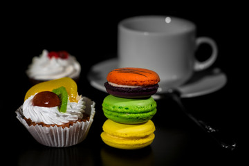 Cake from white cream and fruits and vivid colored macaroons in stack spot lighted on black background with cup stand on saucer/shallow depth of field