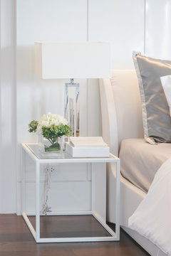 Flower jar, book and reading lamp on white frame bedside table next to bed in modern interior bedroom