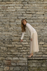 Fototapeta na wymiar Caucasian white female model and brick stone. Beautiful girl, long red hair, beige skirt and cardigan. Woman standing on the stairs in old town, limestone walls background