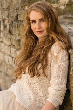 Caucasian white female model portrait, brick stone. Beautiful girl, long red hair, beige skirt and cardigan. Woman siting on the stairs in old town, limestone walls background