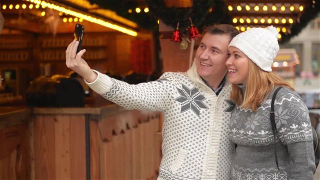 Technology and People concept. Happy Couple of Tourists in Warm Clothes Taking Selfie with Smartphone on the Christmas Market. Young Family Laughing on the Xmas Fair