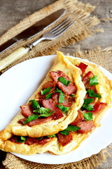 Cooked bacon omelet. Homemade spicy omelet with fried sliced bacon and parsley on a white plate, fork, knife on old wooden table. Very delicious eggs breakfast idea. Rustic style. Closeup
